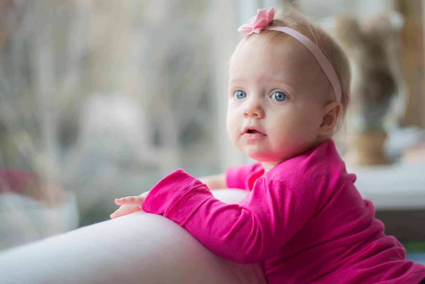 Toddler girl in bright pink shirt resting arms on couch while looking out window