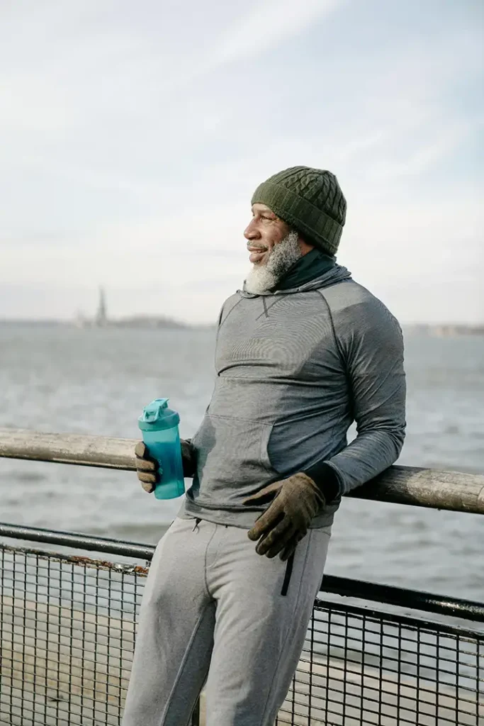 Mature man in activewear stands by the water, holding a tumbler and smiling