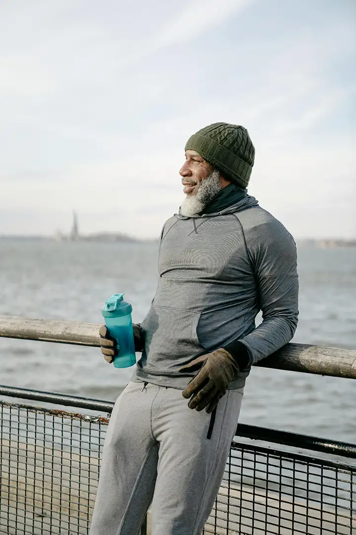 Mature man in activewear stands by the water, holding a tumbler and smiling