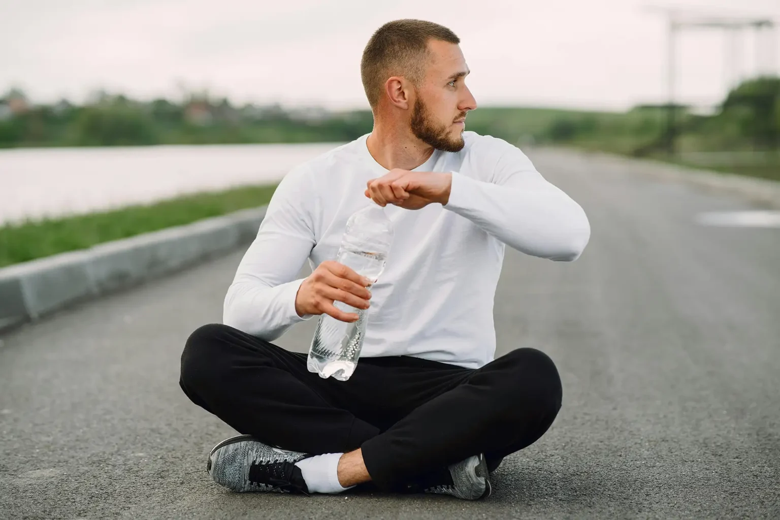 Man seated on an empty road holding a water bottle, focusing on healthy lifestyle