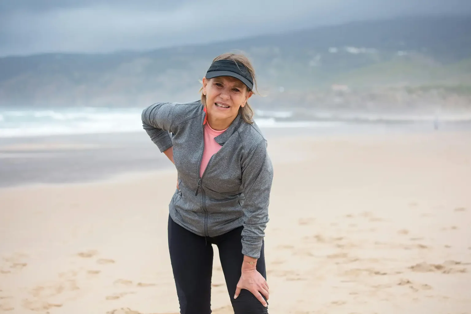 Woman on beach with chronic and joint pain clutching back and knee
