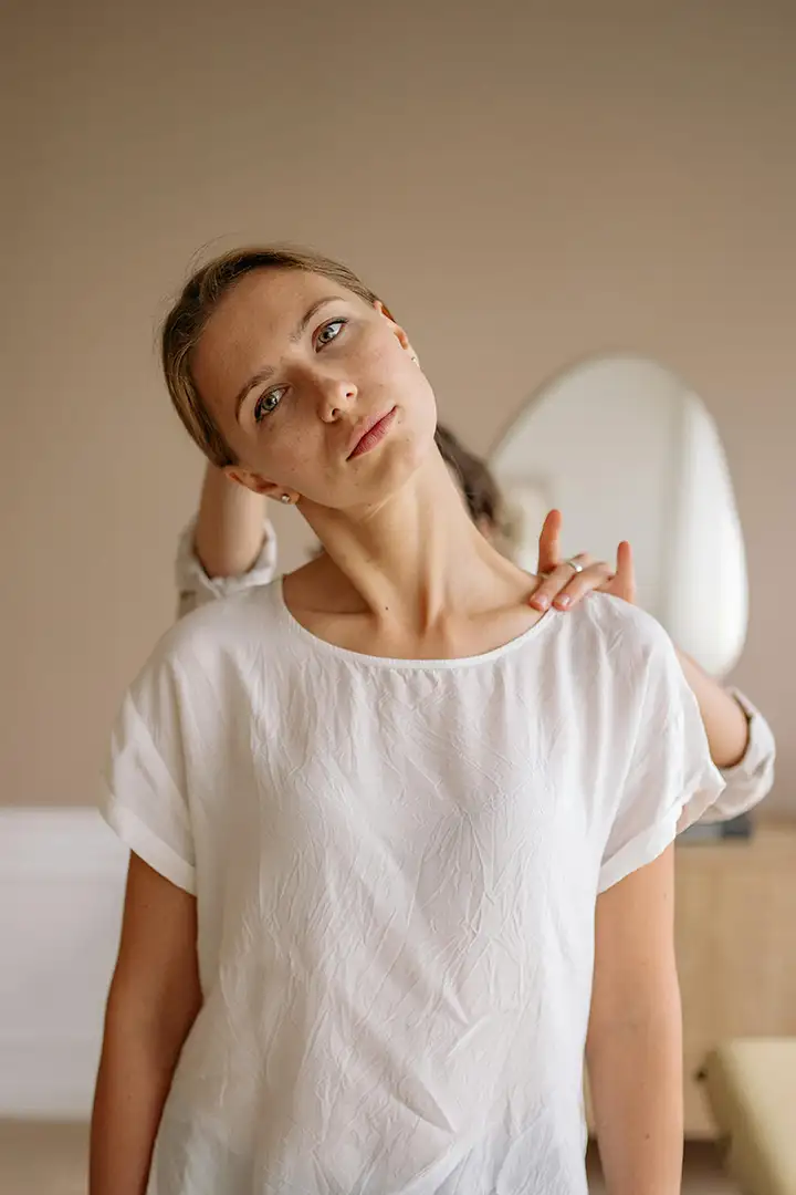 A woman in a white shirt receives a gentle neck stretch from a therapist indoors